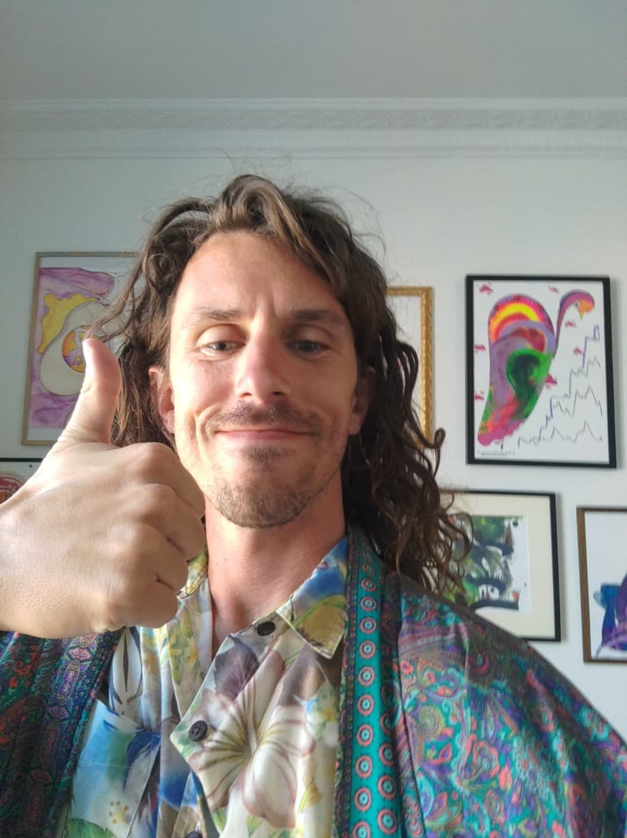 Cole smiling and giving a thumbs up in a colorful floral shirt, in front of a wall of bright framed artwork
