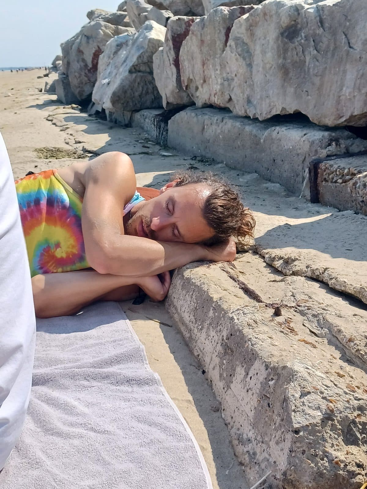Cole asleep on a beach, head resting on his arms against a concrete ledge in front of a row of boulders
